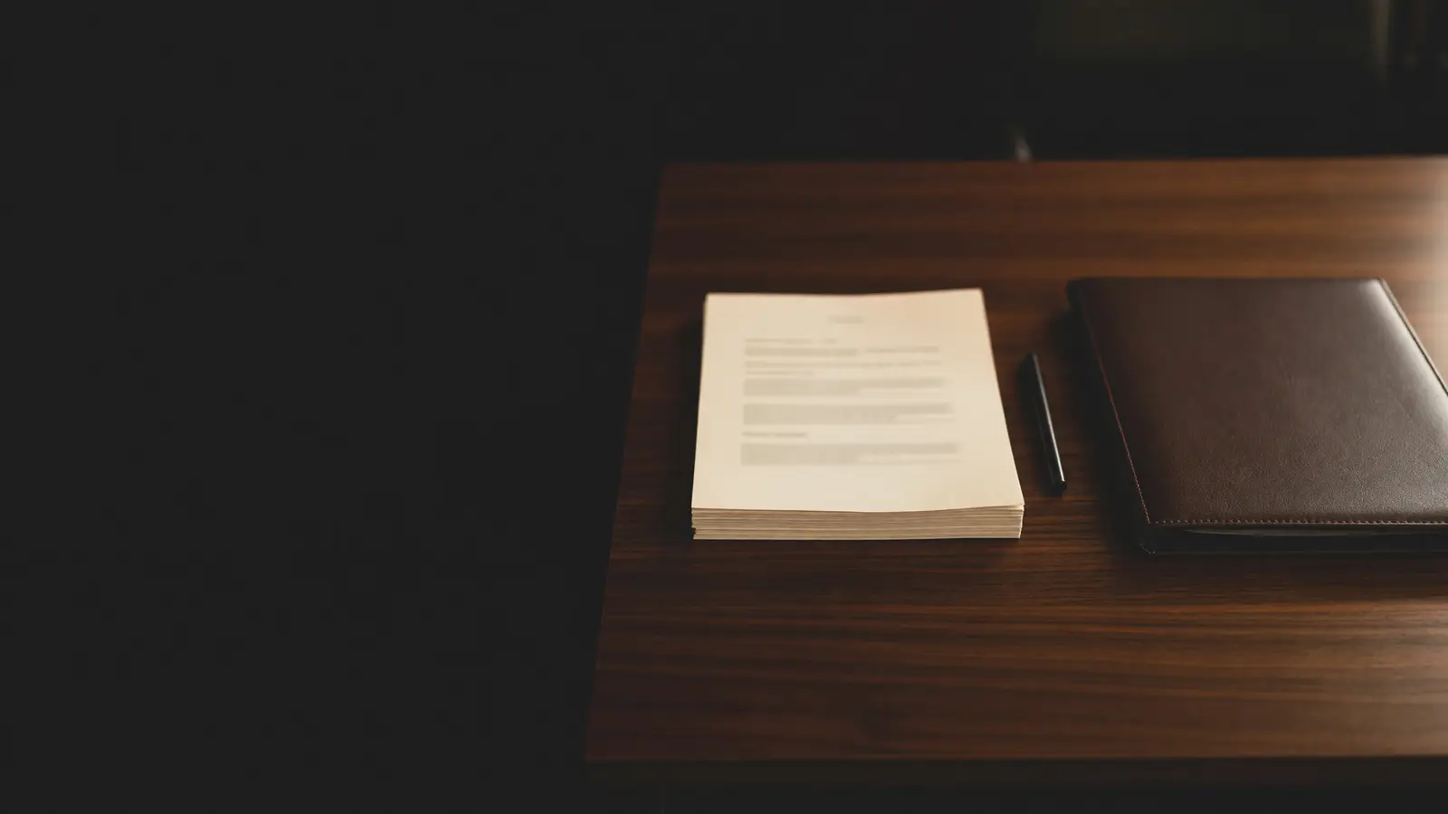 Stacked LLC formation papers, a black pen and a closed leather portfolio on a dark walnut desk in warm light